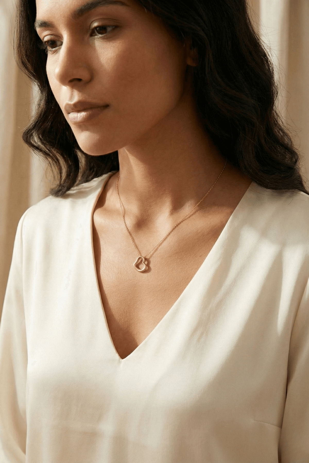 Kadın, 14 ayar altın kalpli kolye takarken, sade beyaz bluz giyiyor. Altın Kolye detayı net görünmekte.